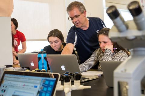 A professor consults with students working on laptops in a lab.